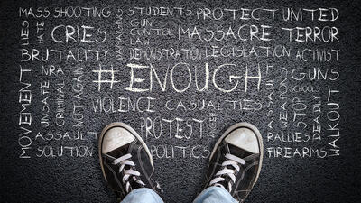 A child's feet with sneakers on them stand in front of words that include protect, students, mass shooting, cries, brutality, activism, walkout and more etched in chalk on the ground. 
